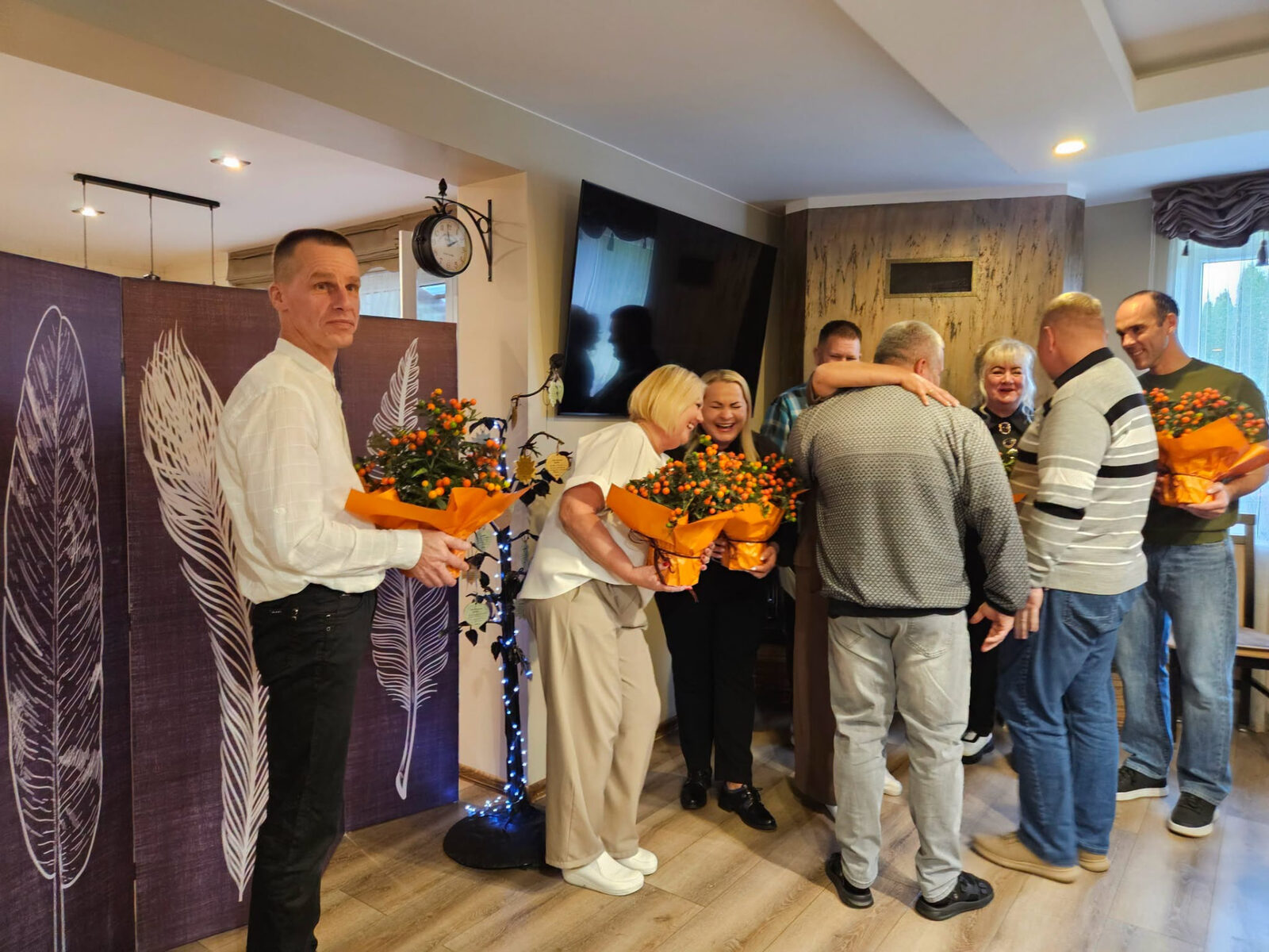 Grupė žmonių su oranžinių gėlių puokštėmis bendrauja ir sveikinasi.